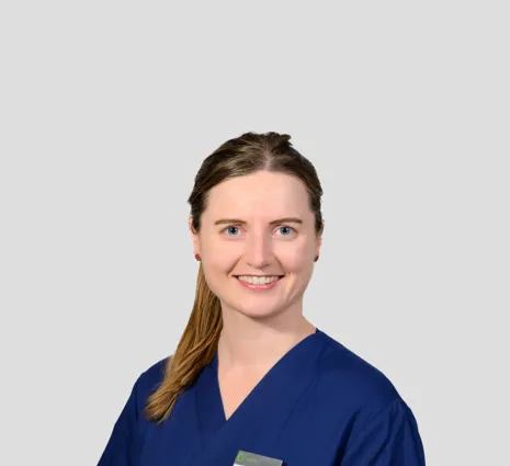 Frau mit langen, braunen Haaren in blauer Arbeitskleidung, lächelt freundlich. Ein Namensschild ist sichtbar. Hintergrund in neutralem Grau.