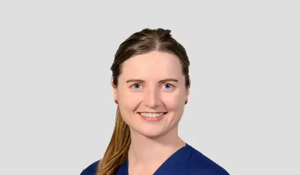 Frau mit langen, braunen Haaren in blauer Arbeitskleidung, lächelt freundlich. Ein Namensschild ist sichtbar. Hintergrund in neutralem Grau.