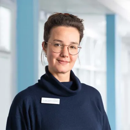 Frau mit Brille und Nasenpiercing trägt einen dunkelblauen Pullover mit hohem Kragen. Sie steht vor einer hellen, modernen Umgebung mit blauen Akzenten und lächelt leicht. Ein Namensschild ist sichtbar.