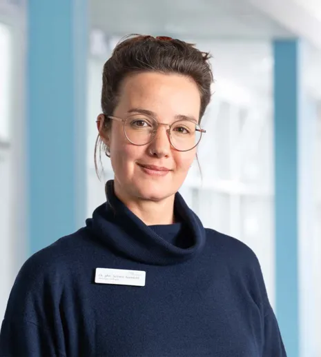 Frau mit Brille und Nasenpiercing trägt einen dunkelblauen Pullover mit hohem Kragen. Sie steht vor einer hellen, modernen Umgebung mit blauen Akzenten und lächelt leicht. Ein Namensschild ist sichtbar.
