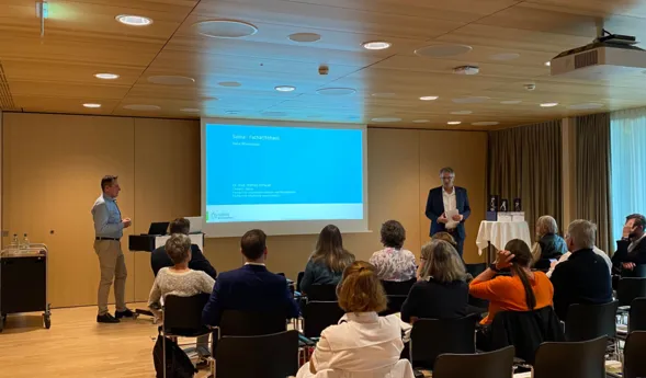 Zwei Männer präsentieren vor einem Publikum in einem modernen Konferenzraum. Der Bildschirm zeigt eine Präsentation mit dem Titel 