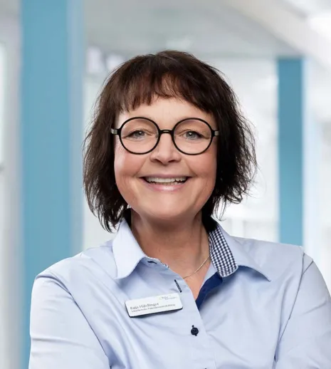 Lächelnde Frau mit kurzen, lockigen Haaren und runden Brillen, trägt ein hellblaues Hemd mit einem karierten Kragen. Im Hintergrund sind helle, moderne Räumlichkeiten sichtbar. Ein Namensschild ist sichtbar.