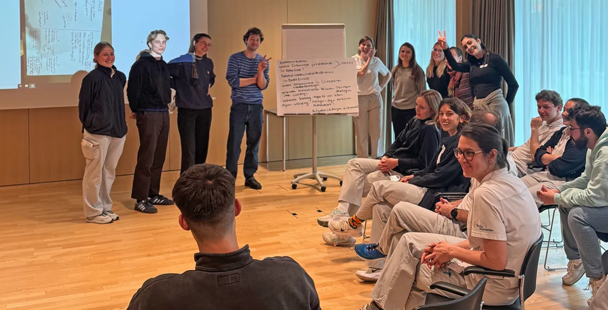 Gruppendiskussion in einem modernen Raum, mit mehreren Personen, die in zwei Gruppen aufgeteilt sind. Eine Gruppe steht vor einer Präsentation, während die andere Gruppe auf Stühlen sitzt und aufmerksam zuhört. An der Wand ist ein Flipchart mit Notizen...