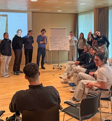 Gruppendiskussion in einem modernen Raum, mit mehreren Personen, die in zwei Gruppen aufgeteilt sind. Eine Gruppe steht vor einer Präsentation, während die andere Gruppe auf Stühlen sitzt und aufmerksam zuhört. An der Wand ist ein Flipchart mit Notizen...