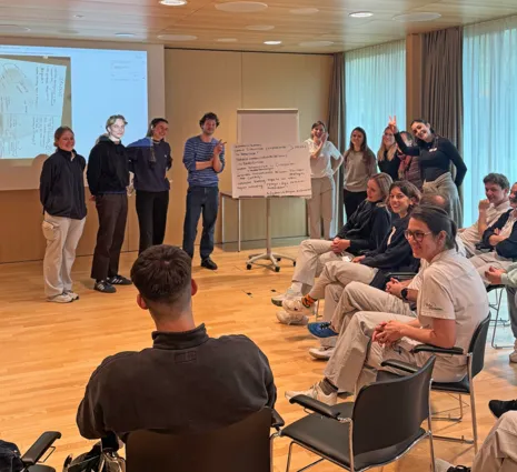 Gruppendiskussion in einem modernen Raum, mit mehreren Personen, die in zwei Gruppen aufgeteilt sind. Eine Gruppe steht vor einer Präsentation, während die andere Gruppe auf Stühlen sitzt und aufmerksam zuhört. An der Wand ist ein Flipchart mit Notizen...