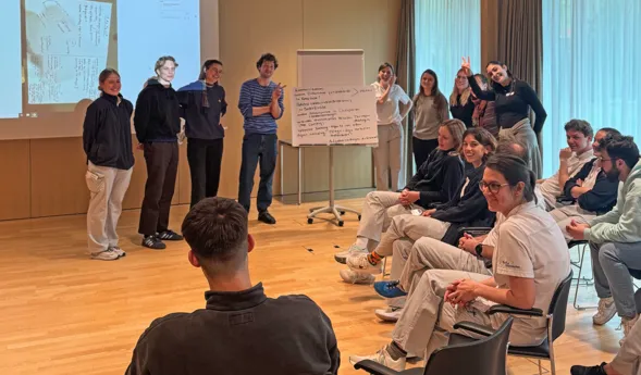 Gruppendiskussion in einem modernen Raum, mit mehreren Personen, die in zwei Gruppen aufgeteilt sind. Eine Gruppe steht vor einer Präsentation, während die andere Gruppe auf Stühlen sitzt und aufmerksam zuhört. An der Wand ist ein Flipchart mit Notizen...