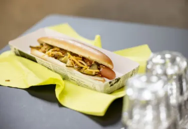 Hotdog in einem Pappbehälter, belegt mit gehackten Gurken, Käse und Zwiebeln, auf einem gelben Serviette, im Hintergrund Glasbehälter.