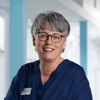 Lächelnde Frau mit grauen, lockigen Haaren und Brille, trägt einen blauen Kittel. Im Hintergrund sind helle, moderne Räumlichkeiten sichtbar. Ein Namensschild ist an ihrer Kleidung befestigt.