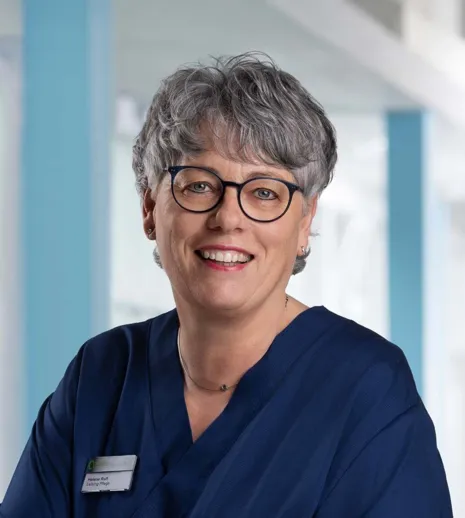 Lächelnde Frau mit grauen, lockigen Haaren und Brille, trägt einen blauen Kittel. Im Hintergrund sind helle, moderne Räumlichkeiten sichtbar. Ein Namensschild ist an ihrer Kleidung befestigt.