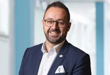 Mann mit Brille und Bart, trägt einen dunkelblauen Anzug mit einem weißen Hemd und einer weißen Einstecktuch, lächelt freundlich vor einem hellen, modernen Hintergrund.