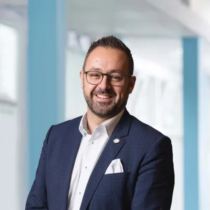 Mann mit Brille und Bart, trägt einen dunkelblauen Anzug mit einem weißen Hemd und einer weißen Einstecktuch, lächelt freundlich vor einem hellen, modernen Hintergrund.