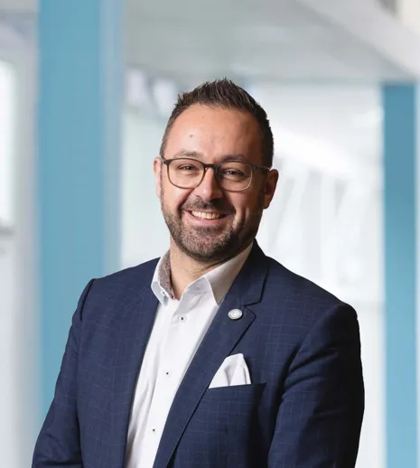 Mann mit Brille und Bart, trägt einen dunkelblauen Anzug mit einem weißen Hemd und einer weißen Einstecktuch, lächelt freundlich vor einem hellen, modernen Hintergrund.