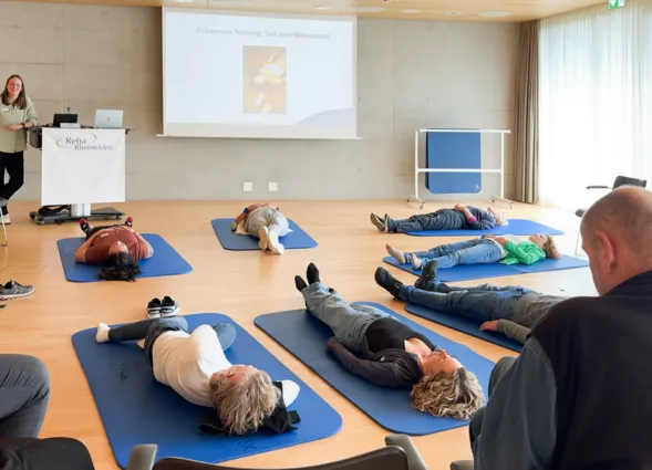 Gruppensitzung im Raum mit Holzfußboden, Teilnehmer liegen auf blauen Matten und praktizieren autogenes Training. Eine Trainerin steht neben einem Präsentationsbildschirm mit dem Titel „Autogenes Training: Zeit zum Mitmachen“. Stühle sind im Hintergrun...