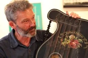 Mann mit grauem Bart hält ein kunstvoll verziertes Zupfinstrument mit Blumenmotiven in der Hand.