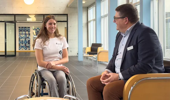 Junge Frau im Rollstuhl und Mann in Anzug sitzen sich in einem modernen Raum gegenüber. Zwischen ihnen steht ein kleiner Tisch mit Zeitschriften. Der Raum hat große Fenster und eine helle, freundliche Atmosphäre.