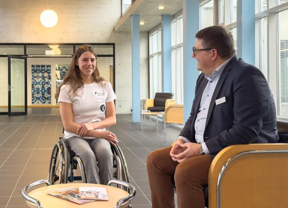 Junge Frau im Rollstuhl und Mann in Anzug sitzen sich in einem modernen Raum gegenüber. Zwischen ihnen steht ein kleiner Tisch mit Zeitschriften. Der Raum hat große Fenster und eine helle, freundliche Atmosphäre.