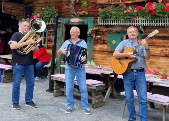 Musikgruppe mit drei Männern auf einer Terrasse vor Holzfassade: Posaune, Akkordeon und Gitarre, im Hintergrund „Insperbach-Alm“ mit Blumenampeln und Bänken.