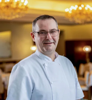 Chef in weißer Kochjacke mit Brille lächelnd im festlich beleuchteten Restaurant, unscharfer Hintergrund mit Tischen und Kronleuchtern.