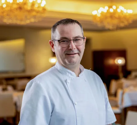 Chef in weißer Kochjacke mit Brille lächelnd im festlich beleuchteten Restaurant, unscharfer Hintergrund mit Tischen und Kronleuchtern.