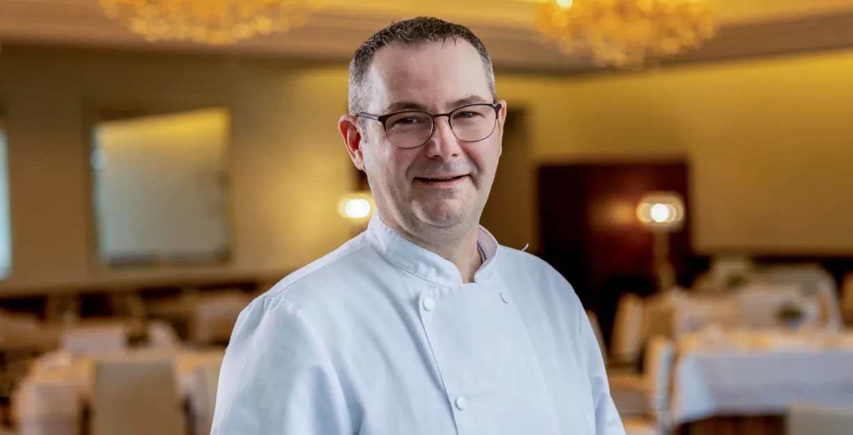 Chef in weißer Kochjacke mit Brille lächelnd im festlich beleuchteten Restaurant, unscharfer Hintergrund mit Tischen und Kronleuchtern.