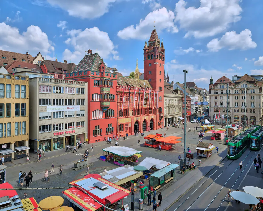 Der imposante Turm, die rote Fassade, und die verspielten Fassadenmalereien machen das Basler Rathaus zu einem Wahrzeichen der Stadt. Davor bietet der Stadtmarkt frisches Obst und Gemüse, Kräuter, Blumen, und andere lokale Erzeugnisse. // The Town hall...