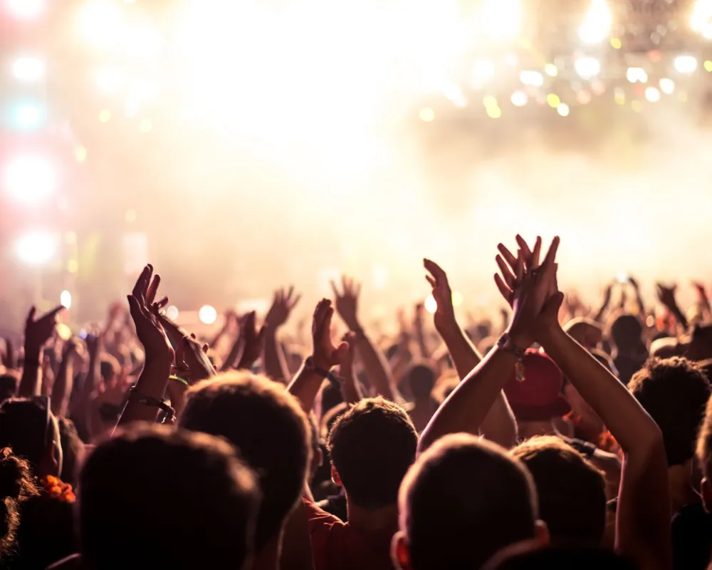 Audience with hands raised at a music festival and lights streaming down from above the stage. Soft focus, blurred movement.