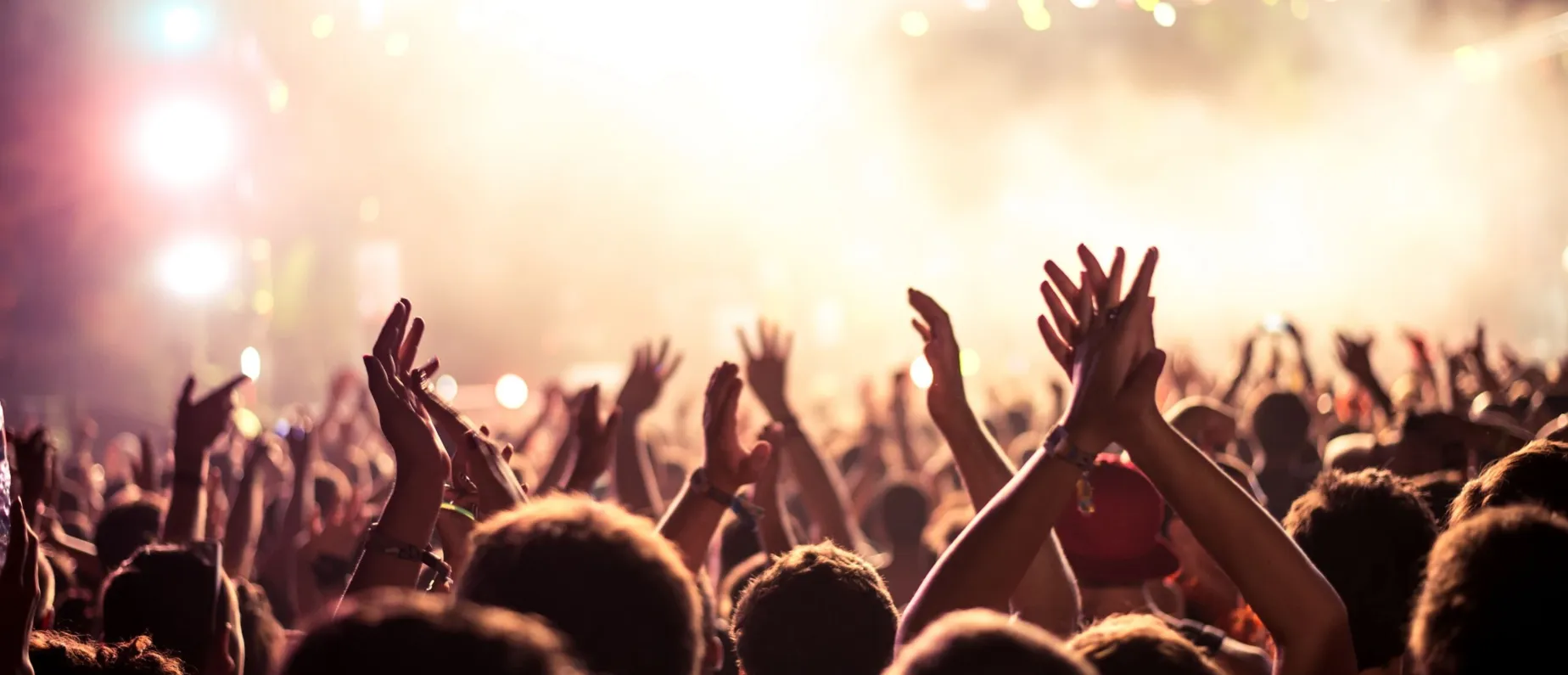 Audience with hands raised at a music festival and lights streaming down from above the stage. Soft focus, blurred movement.