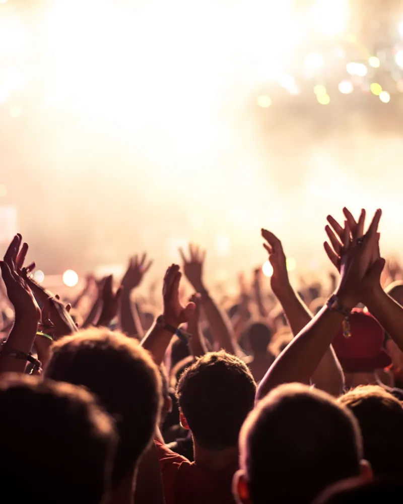 Audience with hands raised at a music festival and lights streaming down from above the stage. Soft focus, blurred movement.