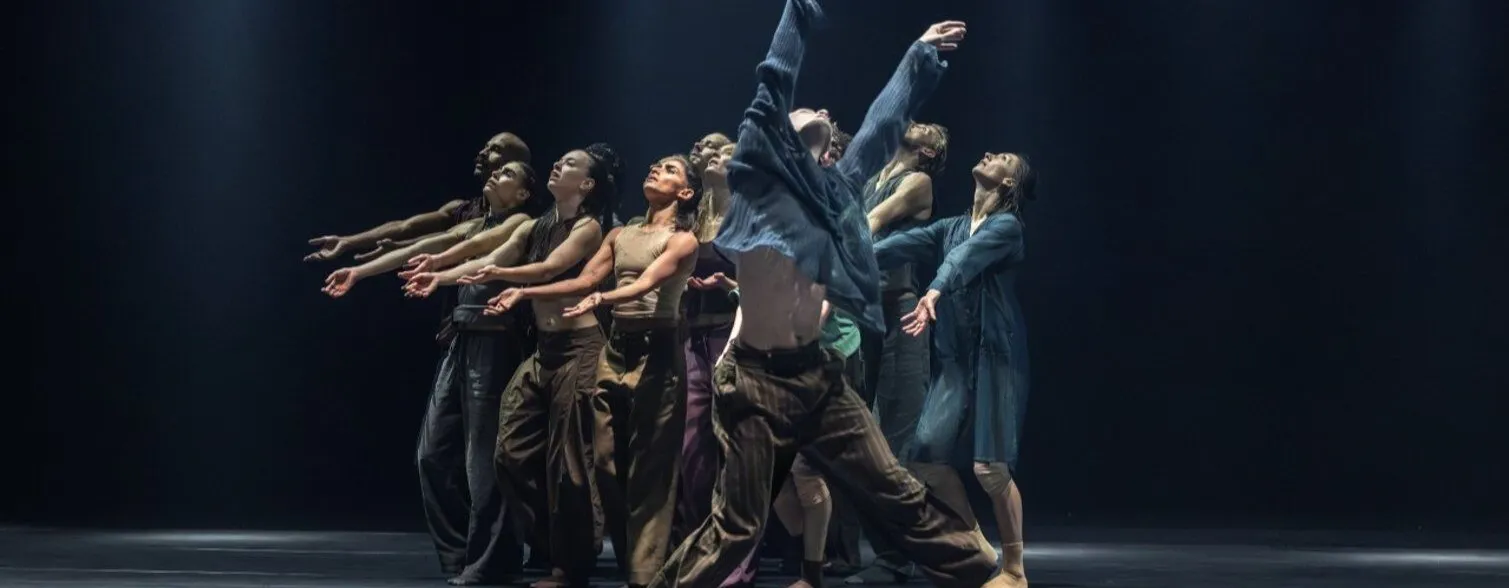 Eine Gruppe von Tänzern in verschiedenen Posen, die in einem schwach beleuchteten Raum agieren. Der Vordergrund zeigt einen Tänzer mit freiem Oberkörper, während die anderen Tänzer hinter ihm in dynamischen Bewegungen stehen. Die Kleidung ist lässig un...