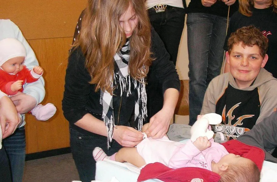 Mädchen mit langen Haaren und Schal wechselt die Windel einer Puppe auf einem Wickeltisch, während andere Kinder und Jugendliche im Hintergrund zuschauen. Eine weitere Puppe wird von einem anderen Kind gehalten.