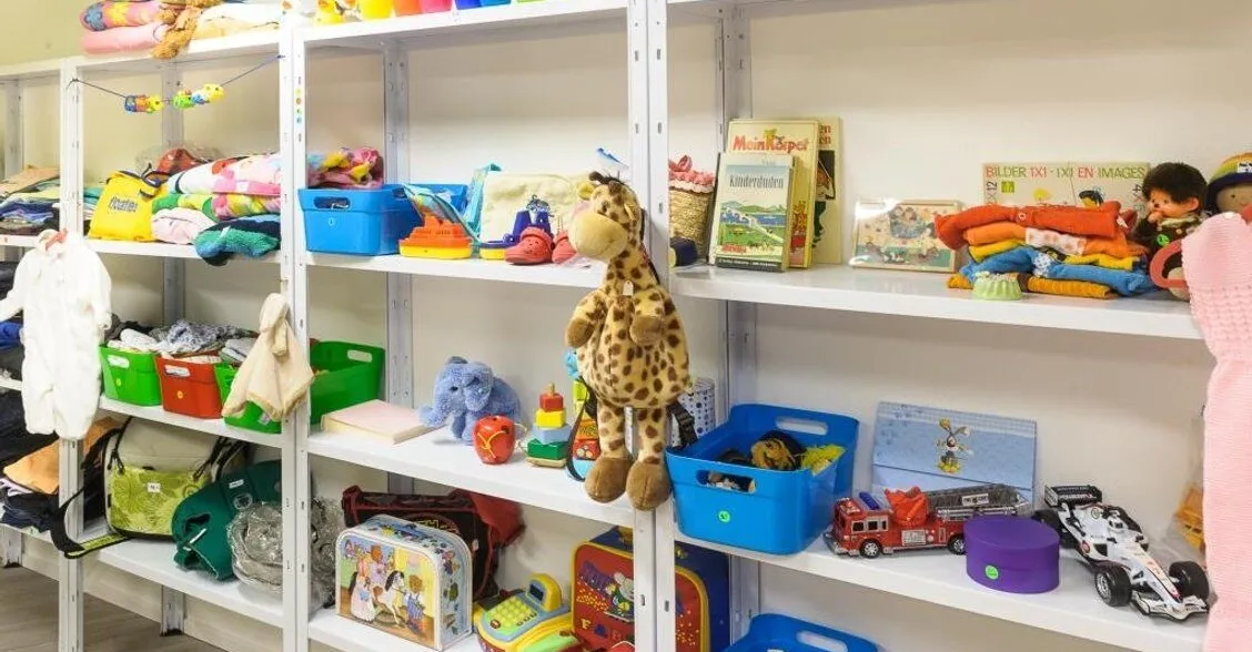 Regale mit bunten Spielsachen, Stofftieren, Büchern und Textilien. Verschiedene Aufbewahrungskisten in Grün und Blau sowie eine Vielzahl von Spielzeugen, darunter ein Plüschgiraffe und Fahrzeuge.