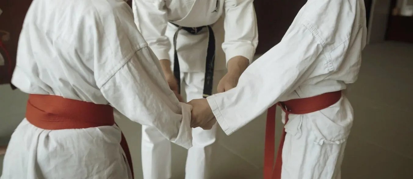 Drei Personen in Karate-Uniformen stehen in einem Dojo. Zwei Kinder mit roten Gürteln halten sich an den Händen, während ein Erwachsener in der Mitte die Hände der Kinder führt. Der Boden ist hell und die Wände sind dunkel.