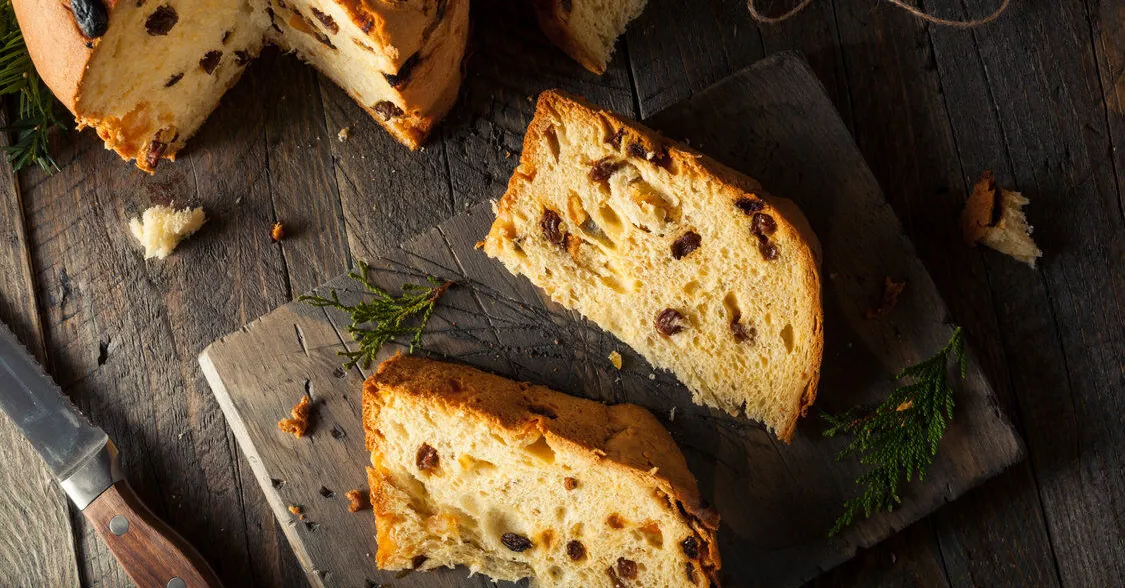 Sliced bread with raisins on a wooden cutting board, accompanied by a knife and scattered crumbs, with green foliage accents.