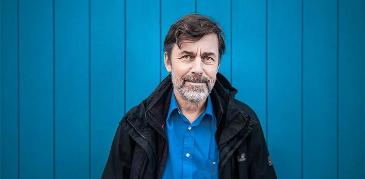 Mann mit Bart und lächelndem Gesicht, trägt eine schwarze Jacke über einem blauen Hemd, vor einer hellblauen Wand.