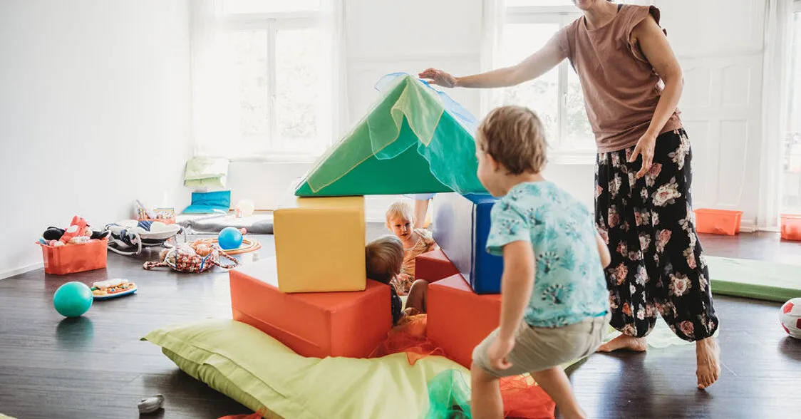 Spielende Kinder und eine Erwachsene in einem hellen Raum mit bunten Schaumstoffblöcken, Tüchern und Spielzeug. Ein Kind versteckt sich in einer Konstruktion aus Blöcken, während ein anderes heranläuft.