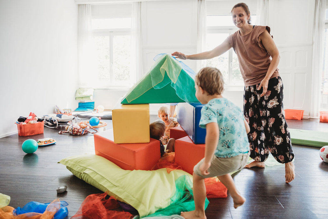 Spielende Kinder und eine Erwachsene in einem hellen Raum mit bunten Schaumstoffblöcken, Tüchern und Spielzeug. Ein Kind versteckt sich in einer Konstruktion aus Blöcken, während ein anderes heranläuft.