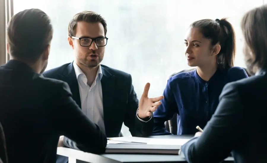 Confident young businessman in eyeglasses holding negotiations meeting with partners in formal wear. Focused diverse employees discussing development strategy or working issues, sitting at table.