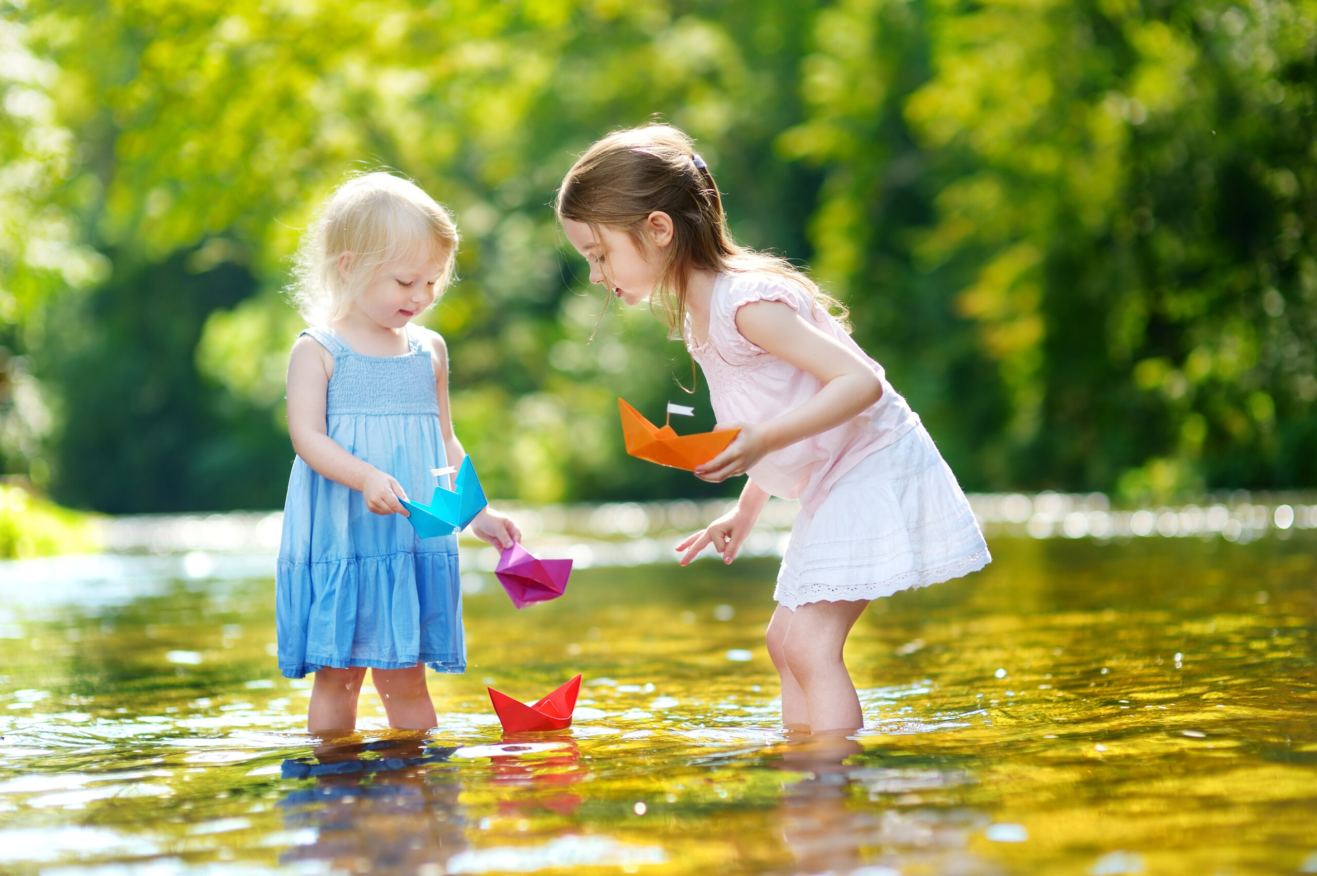 Two little sisters playing with paper boats by a river on warm and sunny summer day