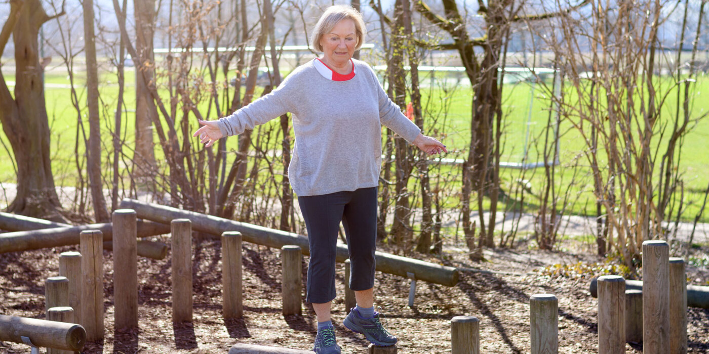 Senior woman balancing on wooden pillar in park, standing with her hands spread