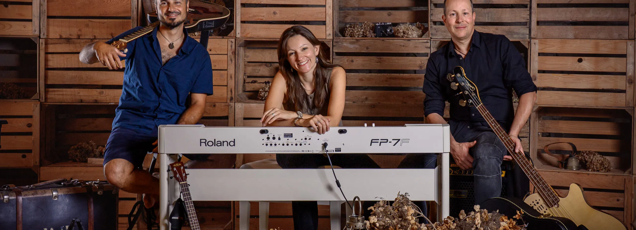 Drei Musiker sitzen in einem rustikalen Raum mit Holzwand, Keyboard, Gitarren, Kontrabass, Schlagzeug und Körben mit Trockenblumen.