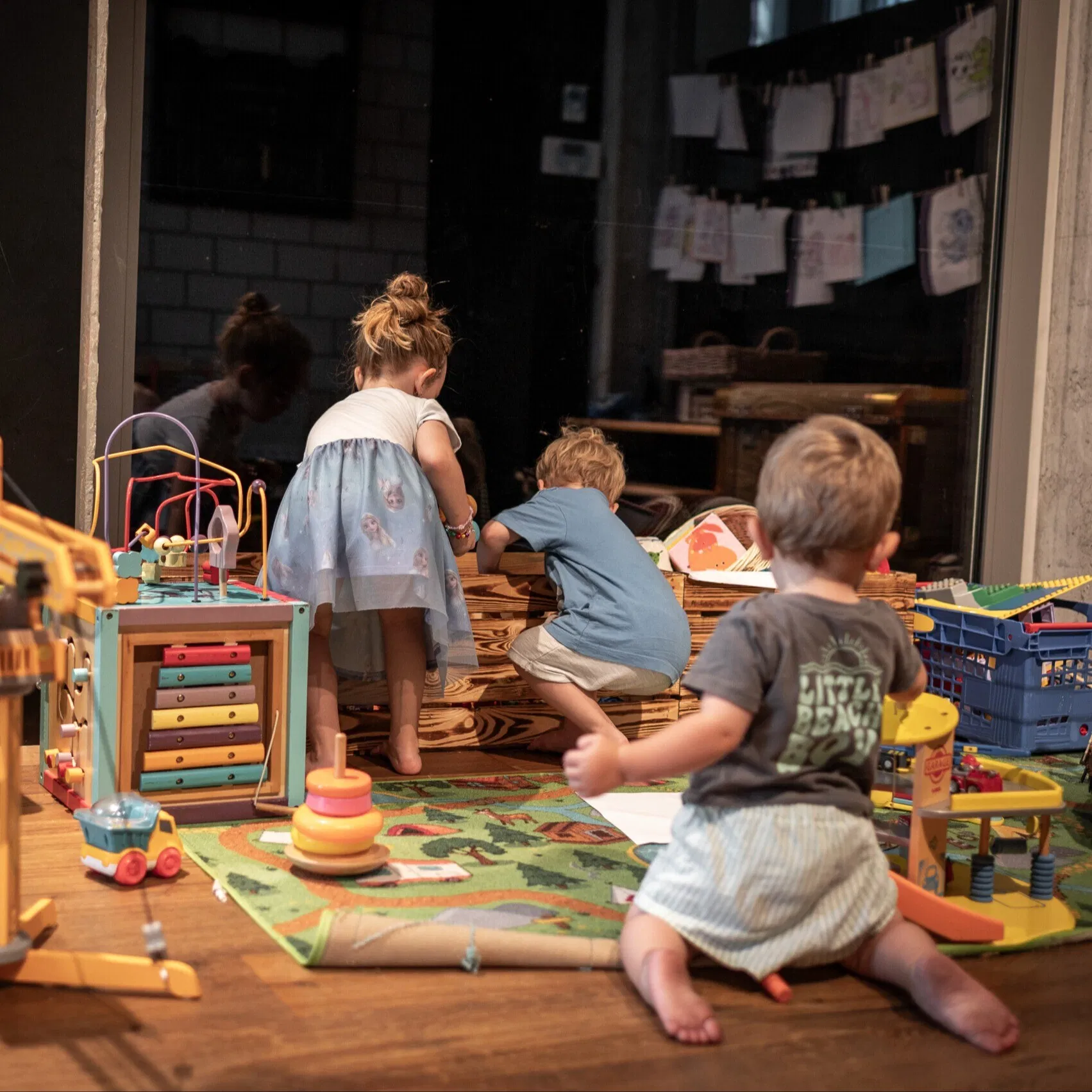 Kinder spielen in einem Spielbereich mit Holzspielzeug, Puppen und Spielmatte; im Hintergrund hängen eine Zielscheibe und Kinderzeichnungen.